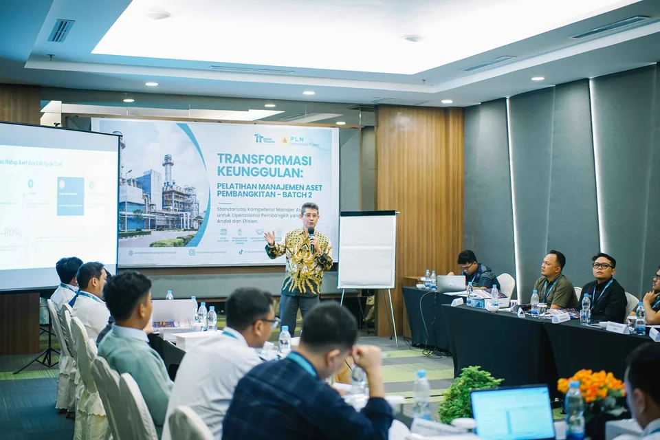 Pembicara berbatik memegang mikrofon di depan sekitar sepuluh peserta di ruang rapat hotel, dengan spanduk Pelatihan Manajemen Aset Pembangkitan Batch 2.
